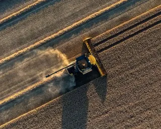 An aerial view of a combine harvester moving through rows of harvested crop, creating a dust trail