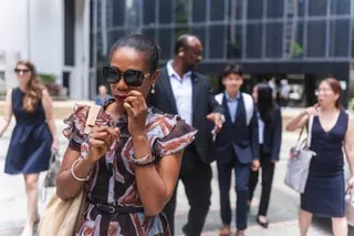 Aysha Bell leaving the court. She is a black woman with her hair tied back and wears large sunglasses and a short-sleeved dress in a red, white and blue geometric pattern. The court building behind her has a modern glass frontage.