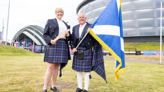 a man and woman in full Highland gear holding a scotland flag and baton outside the Hydro venue