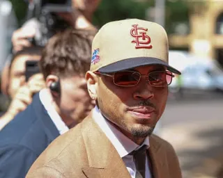 Chris Brown in a tan suit and baseball cap with tinted sunglasses looks toward the camera