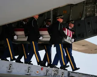 Soldiers in dress uniforms with yellow stripes down pants carry a coffin out of an aircraft.