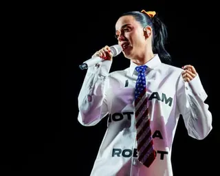 Katy Perry in a white shirt and sparkly tie sings into a microphone on a dark stage