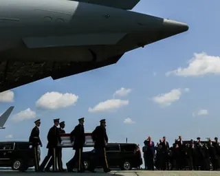 line of people standing outside salute as soldiers carry casket next to plane