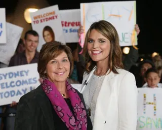 two women standing together smiling, as people behind them hold signs saying 'sydney welcomes today' and 'welcome to sydney'