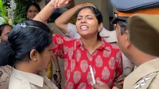 Somaya Korde is surrounded by the police at a protest. Wearing a red shirt with white leaves pictured on it, she holds her arm up in defiance and has her mouth slightly open as if she's shouting.