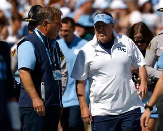 Michael Lombardi, wearing a blue vest, stands on a college football game sideline with Bill Belichick, wearing a white polo shirt