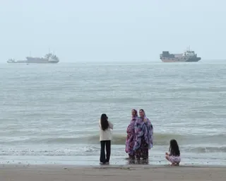 Four people stand at the water's edge on a beach in the Strait of Hormuz, with cargo ships visible in the hazy distance