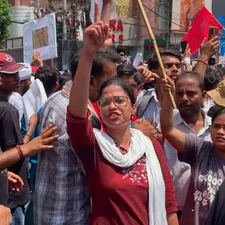 Varuni Poorva, an aspiring professor, raises her hand during a protest. She wears a red top with flowers embroidered on it in white. She has a white scarf over her shoulders and is surrounded by other protesters.