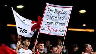 A crowd of people. One of the people is holding a large banner which reads: 'The Mayo Curse is here to stay Sam's returning to Tyrone today'.