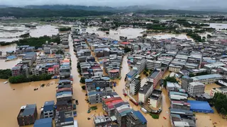 Aerial view of homes and buildings submerged in brown flood waters in Hengzhou, Nanning, China with a green mountain range and clody skies in the background