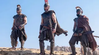 Three soldiers from the film The Odyssey standing in a row looking into the distance with a blue sky behind them