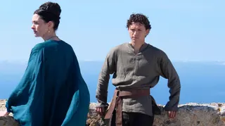 Tom Holland and Anne Hathaway in a film still from the film The Odyssey. Tom Holland is leaning against a wall with a scenic greek sea view  behind him. He is wearing a dark olive green shirt and dark trousers. Anne hathaway is looking to the left and wearing a flowing flowing blue dress