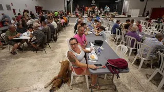 Wide shot of a shelter in Spain with three long plastic tables and plastic garden chairs around them. People evacuated from their homes are sitting at the tables eating and talking