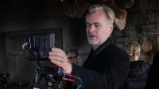 Christopher Nolan in the centre of the photo and is looking through an Imax camera. He is wearing a black jacket.
