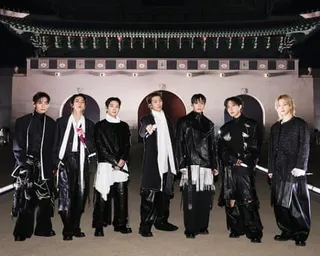 Seven members of BTS in black and white outfits stand in a line before a traditional architectural backdrop