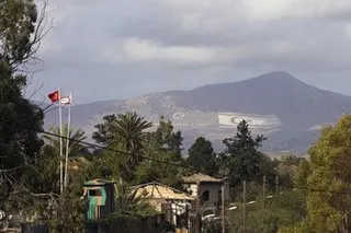 塞浦路斯联合国驻UNFICYP维持和平部队缓冲区景色，前景中旗杆上飘扬着土耳其国旗和联合国旗帜，背景中可以看到一座山。