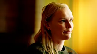 A close-up portrait of Alex looking serious, with pursed lips. Her face is turned to the side and she is looking towards the right of the frame. She has blonde hair and wears a dark top and small earring. The background is softly blurred in shades of yellow, gold, and dark brown.