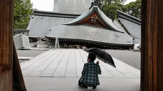 A woman with an umbrella squats in front of a collapsed shrine