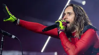 Jared Leto stands under concert lighting, singing into a handheld microphone. He has long hair and a beard and is dressed in a red, sheer and fitted top with neon green and pink gloves. One arm is stretched outward toward the audience while the other holds the microphone close to his mouth. Bright white stage lights form diagonal lines in the background.


CLARKSTON, MICHIGAN - AUGUST 06: Actor/Singer Jared Leto of Thirty Seconds To Mars performs during their Seasons World Tour 2024 at Pine Knob