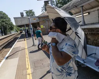 A man with glasses holds an umbrella and wipes his face with a cloth on a train platform