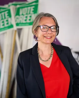 Geraldine Coggins stands in front of signs saying Vote Green. She is 50 with short greying hair and glasses, and wears a bright red top under a dark jacket with a chunky chain-style necklace.