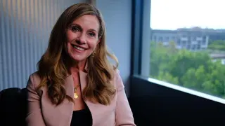 Julie Inman Grant in a pale pink jacket and black blouse smiles as she looks at the camera.  She is sitting next to a window.