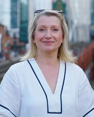 Sian Astley stands with tall buildings of Manchester behind her. She is in her 50s and has shoulder-length straight blond hair; she wears a loose white tunic-style linen shirt.