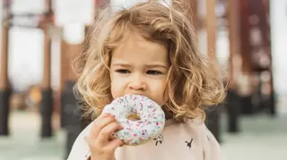 圖片顯示一名年幼兒童在戶外手持一個大甜甜圈，甜甜圈上覆有白色糖霜和彩色糖粒。兒童正咬下一口甜甜圈，而甜甜圈佔據了前景的大部分空間。背景可見模糊的結構元素，例如柱子或樑架，營造出淺景深效果。柔和的自然光突顯了兒童的捲髮，以及衣著和甜甜圈糖霜的柔和色調。
