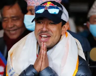 Nirmal Purja in a cap and white scarf holds his hands together in a prayer gesture, smiling