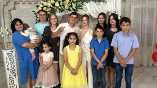 Twelve family members smile at the camera while what appears to be a bride and groom hold up their traditional wedding ring fingers