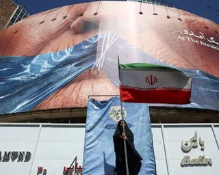 A woman holds an Iranian flag beneath a billboard in Tehran depicting Donald Trump’s mouth sewn shut with a ribbon in the shape of the strait of Hormuz