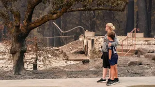 A woman dressed in black capris, grey jumper and black trousers wraps her arm around a young boy in black shorts, black top and black trainers, as both survey the damage of a burnt tree and rubble of a building