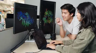 Two people working together at a desk in what appears to be a laboratory or research office environment. Large computer monitors display colorful three-dimensional molecular or protein structure models against dark backgrounds, while a laptop on the desk shows lines of code or data. One person is seated using the laptop, and another is leaning toward the screens as both focus on the scientific visualizations. Laboratory shelving and equipment are visible in the background, suggesting a biomedica