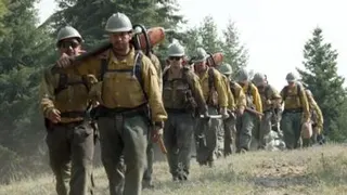 A line of men in firefighting gear walk through an opening in a forest towards the camera lens.