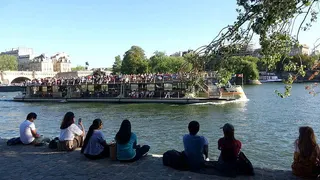 巴黎塞納河上滿載遊客的觀光船往來穿梭，岸邊樹蔭下也坐滿了乘涼賞景的民眾，顯示巴黎...