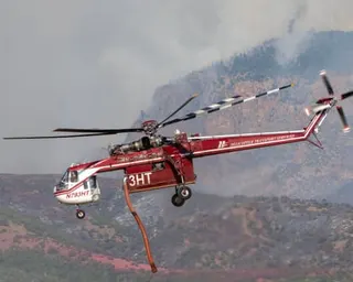 Red and white helicopter with hose trailing, above hills.