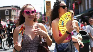 Two young women in sunglasses walking in the sun with brightly coloured fans near their faces. One of them is wearing bright pink heart shaped sunglasses