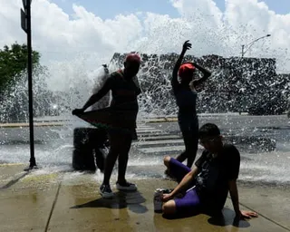 two people stand as water sprays from an open fire hydrant while another person sits on the ground