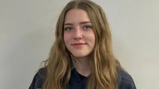 An image showing Keegan Radomski. She has long slightly wavy blonde hair, blue eyes and is wearing a black shirt. Behind her is a plain, white backdrop.
