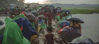 雨中，移民儿童撑着伞，沿着泥泞的小路行走，路两旁搭着用油布临时搭建的遮雨棚。