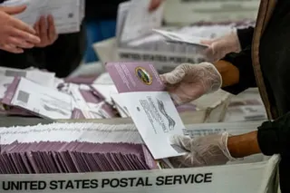 Election workers wearing gloves sort mail-in ballot envelopes in United States Postal Service bins.