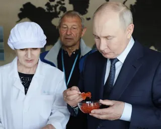 Vladimir Putin samples caviar from a small dish, with a woman in a white overcoat and cap standing next to him