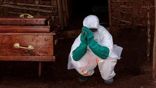 A health worker in full PPE - white suit and green rubber gloves - crouched on the right of a wooden coffin with gold handles