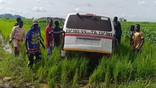 A group of men in Nigeria gather around a white vehicle that is stuck in a muddy rural area surrounded by long grass