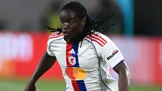 Tabitha Chawinga visible from waist up while in action wearing a white Lyon shirt with red and blue detail running down its centre. She puffs out her cheeks as she runs during a football match, with her many braids flapping in all directions behind and to the side of her head