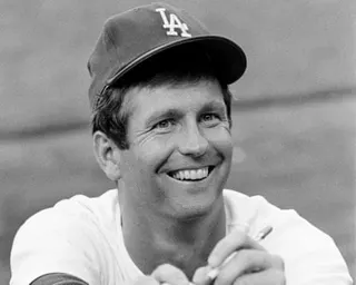 Tommy John smiles while holding a pen, signing an autograph in his Dodgers cap