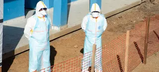 两名身穿防护装备的医务人员站在刚果民主共和国布尼亚市一座正在建设中的蓝色大楼外，周围设有橙色安全围栏。