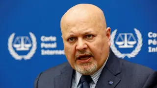 International Criminal Court Prosecutor Karim Khan speaks during an interview while sitting in front of a blue sign with blurry white writing and the symbol of the ICC - the scales of justice surrounded by a laurel wreath.