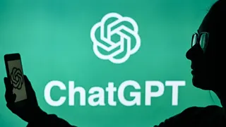 A woman holds a cell phone in front of a computer screen displaying the ChatGPT logo
