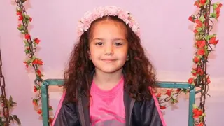 Hind Rajab smiles at the camera. She has long, brown wavy hair and is wearing a pink flower crown on her head. She also has a pink T-shirt and black zip-up on. She is sitting on a chair adorned with garlands of red flowers.